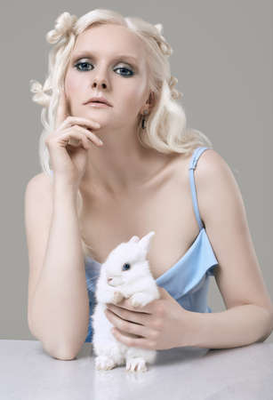 Fashion portrait of albino blond girl with white skin and blue eyes in elegant dress posing with cute little rabbit on gray background in studioの写真素材