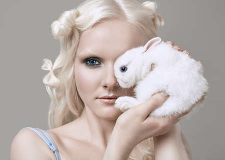 Fashion portrait of albino blond girl with white skin and blue eyes in elegant dress posing with cute little rabbit on gray background in studioの写真素材