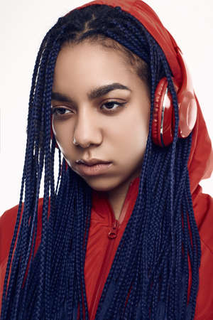 Portrait of beautiful african brunette teenage girl with dreadlocks wearing a red windbreaker listening music via wireless headphones isolated on studio backgroundの写真素材