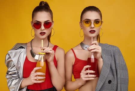 Portrait of two elegant glamor hipster twin girls in fashion red top, black shorts and sunglasses with cocktail drink in studioの写真素材