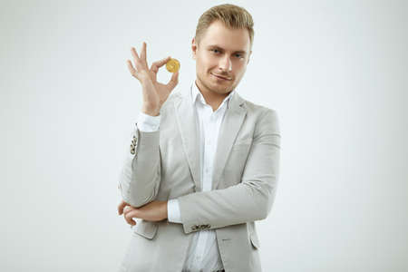 Portrait of a handsome blonde man model in a fashion gray suit holds a Bitcoin isolated on white...