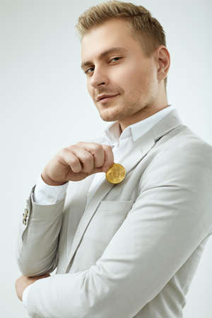 Portrait of a handsome blonde man model in a fashion gray suit holds a Bitcoin isolated on white background in studioの写真素材