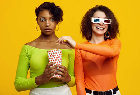Portrait of two young beautiful hipster girls in trendy colorful summer clothes eating popcorn and watching a stereo movie on yellow background.の写真素材