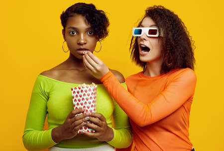 Portrait of two young beautiful hipster girls in trendy colorful summer clothes eating popcorn and watching a stereo movie on yellow background.の写真素材