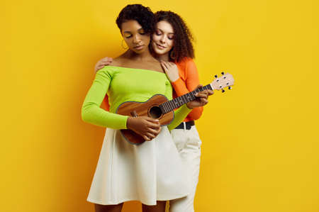 Portrait of two young beautiful hipster girls in trendy colorful summer clothes playing the small guitar on yellow background. Crazy positive bright teensの写真素材