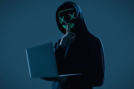 Portrait of an anonymous man in a black hoodie and neon mask hacking into a computer. Studio shot.