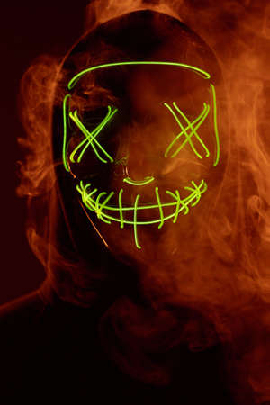 Close-up portrait of an anonymous man hiding his face behind a scary neon mask in a colored smoke....