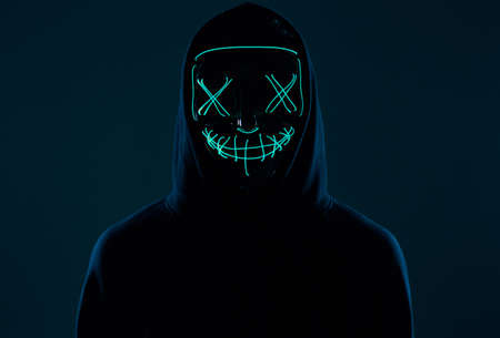 Portrait of an anonymous man in a black hoodie hiding his face behind a scary neon mask. Studio...