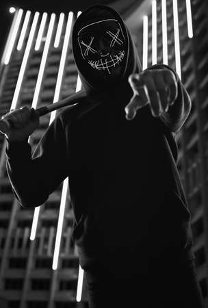 Portrait of an anonymous criminal man with baseball bat in a black hoodie hiding his face behind a...