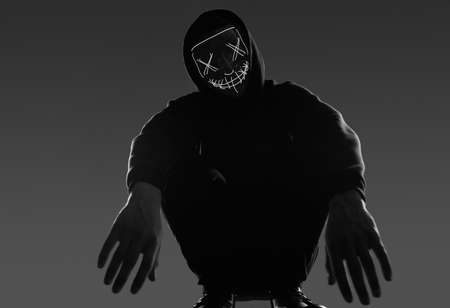 Portrait of an anonymous man in a black hoodie hiding his face behind a scary neon mask. Studio...