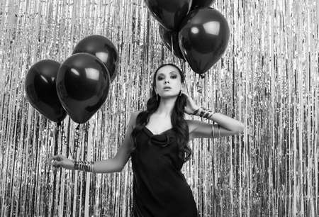 Portrait of gorgeous elegant hispanic brunette woman in luxurious black dress holds balloons and posing on silver shiny tinsel background. Studio shot.の写真素材