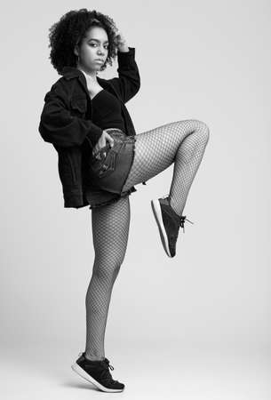Sensual portrait of glamor elegant black hipster teenage model with curly hair in jeans shorts and stockings posing on gray background in studioの写真素材
