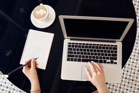 Close-up top view of female hands working on laptop with blank black screen standing on desk in cafe...