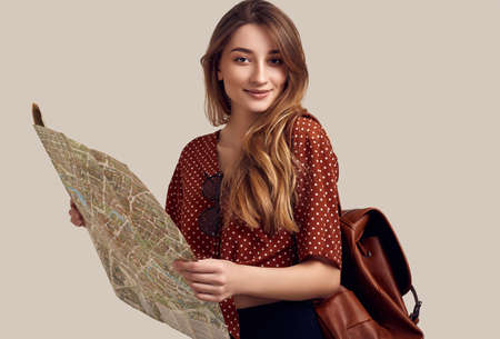 Travel concept. Dreaming about journey. Studio portrait of pretty young hipster woman with backpack holding map. Isolated on gray backgroundの写真素材