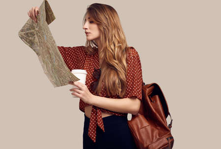 Travel concept. Dreaming about journey. Studio portrait of pretty young hipster woman with backpack holding map. Isolated on gray backgroundの写真素材