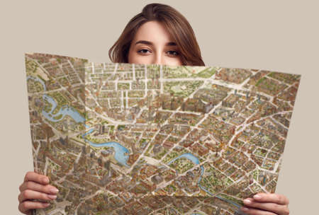 Travel concept. Dreaming about journey. Studio portrait of pretty young hipster woman with backpack holding map. Isolated on gray backgroundの写真素材