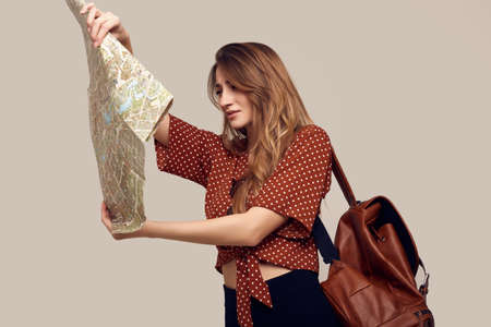 Travel concept. Dreaming about journey. Studio portrait of pretty young hipster woman with backpack holding map. Isolated on gray backgroundの写真素材