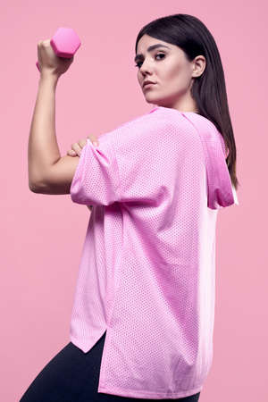Portrait of a gorgeous body positive Latin woman in a pink sports hoodie exercising with dumbbells on a vivid background in the studioの写真素材