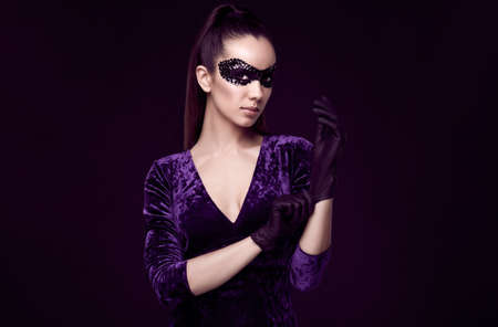 Portrait of charming elegant brunette woman in beautiful purple dress and sequins mask glove up on black background in studioの写真素材