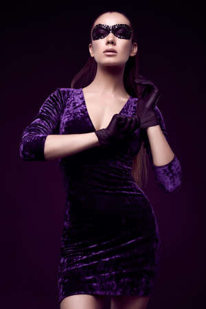 Portrait of charming elegant brunette woman in beautiful purple dress and sequins mask glove up on black background in studioの写真素材