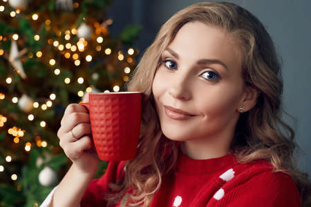 Portrait of gorgeous blonde woman in red sweater chilling under a plaid with cup of coffee in the...