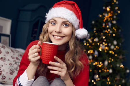 Portrait of gorgeous blonde woman in red sweater and Santa hat with cup of coffee posing in the Christmas decorated interior.の写真素材