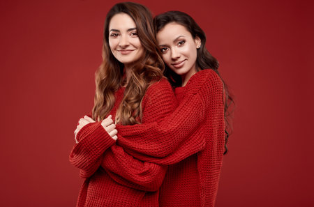 Portrait of two pretty crazy sisters best friends twins in fashion cozy winter sweater posing on red background in studioの写真素材