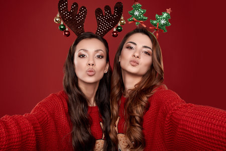 Portrait of two pretty crazy best friends in fashion cozy winter sweater with Christmas attributes taking selfie on red backgroundの写真素材