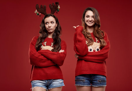 Portrait of two pretty crazy best friends in fashion cozy winter sweater with Christmas attributes posing on red background in studioの写真素材