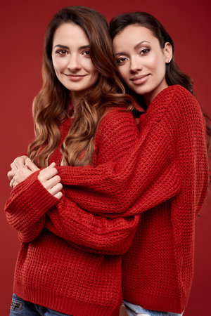 Portrait of two pretty crazy sisters best friends twins in fashion cozy winter sweater posing on red background in studioの写真素材