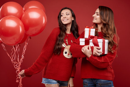 Portrait of two pretty crazy sisters best friends twins in fashion cozy winter sweater posing on red background in studioの写真素材