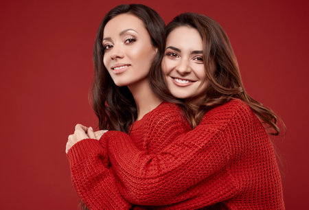 Portrait of two pretty crazy sisters best friends twins in fashion cozy winter sweater posing on red background in studioの写真素材