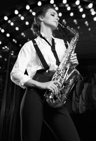 Portrait of gorgeous brunette model woman in fashionable formal suit with saxophone playing on restaurant stage spotlights background.の写真素材