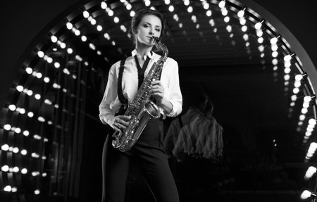 Portrait of gorgeous brunette model woman in fashionable formal suit with saxophone playing on restaurant stage spotlights background.の写真素材