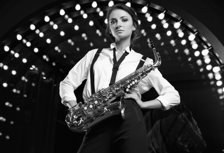 Portrait of gorgeous brunette model woman in fashionable formal suit with saxophone playing on restaurant stage spotlights background.の写真素材