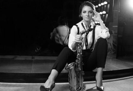 Portrait of gorgeous brunette model woman in fashionable formal suit with saxophone playing on restaurant stage spotlights background.の写真素材