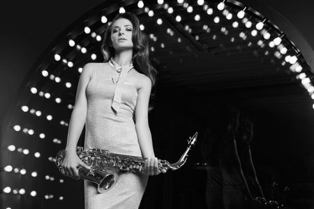 Portrait of gorgeous brunette model woman in elegant dress with saxophone playing on restaurant stage spotlights background.の写真素材