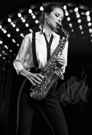 Portrait of gorgeous brunette model woman in fashionable formal suit with saxophone playing on restaurant stage spotlights background.の写真素材