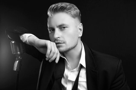 Portrait of handsome blond man singer in elegant tuxedo and bow tie posing with vintage microphone on black studio backgroundの写真素材