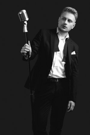 Portrait of handsome blond man singer in elegant tuxedo and bow tie posing with vintage microphone on black studio backgroundの写真素材