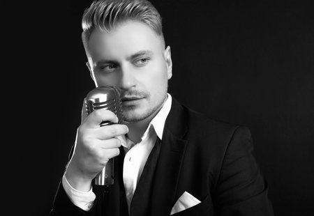 Portrait of handsome blond man singer in elegant tuxedo and bow tie posing with vintage microphone on black studio backgroundの写真素材
