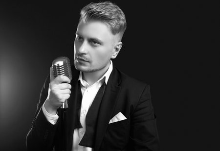 Portrait of handsome blond man singer in elegant tuxedo and bow tie posing with vintage microphone on black studio backgroundの写真素材