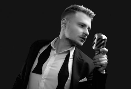 Portrait of handsome blond man singer in elegant tuxedo and bow tie posing with vintage microphone on black studio backgroundの写真素材