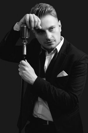 Portrait of handsome blond man singer in elegant tuxedo and bow tie posing with vintage microphone on black studio backgroundの写真素材