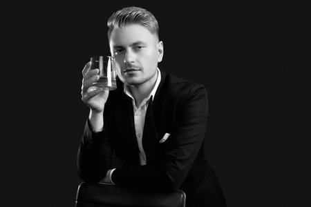 Portrait of handsome blond man in elegant tuxedo and bow tie posing with glass of whiskey on black studio backgroundの写真素材