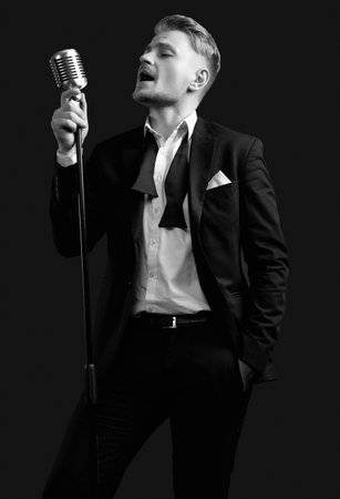 Portrait of handsome blond man singer in elegant tuxedo and bow tie posing with vintage microphone on black studio backgroundの写真素材