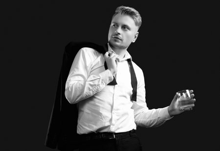 Portrait of handsome blond man in elegant tuxedo and bow tie posing with glass of whiskey on black studio backgroundの写真素材