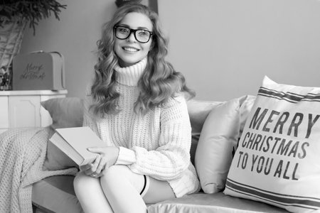 Portrait of gorgeous blonde woman in white woolen sweater and glasses reading book in light cozy decorated interior.の写真素材