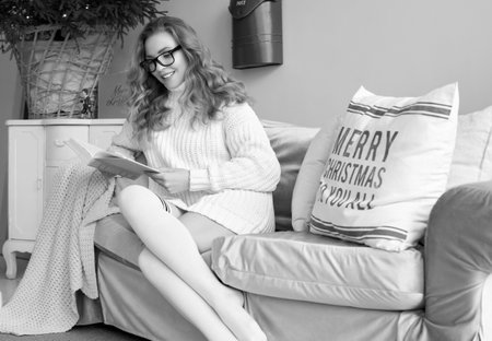 Portrait of gorgeous blonde woman in white woolen sweater and glasses reading book in light cozy decorated interior.の写真素材