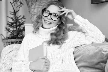 Portrait of gorgeous blonde woman in white woolen sweater and glasses reading book in light cozy decorated interior.の写真素材